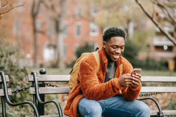 Man in oranje jas op bankje buiten zit op zijn telefoon