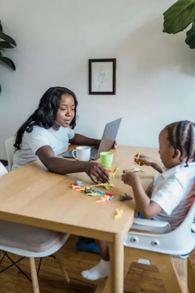 Vrouw achter laptop, kind zit aan tafel te spelen met speelgoed