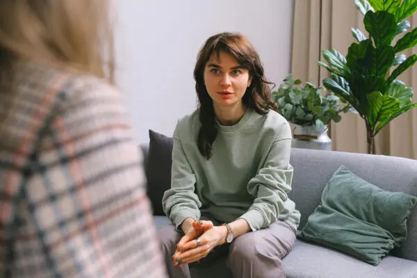 Dame in gesprek met een ander persoon, ze zit op de bank en heeft haar handen in elkaar geslagen